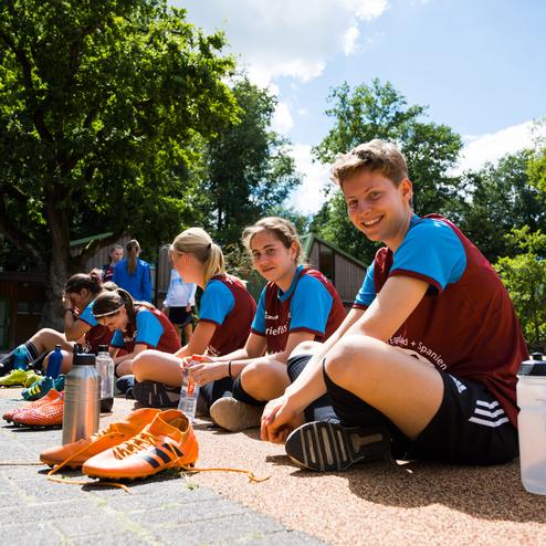 Gruppensitzung junger Fu&szlig;ballspielerinnen auf einem Platz, einige binden die Schuhe, andere trinken Wasser.