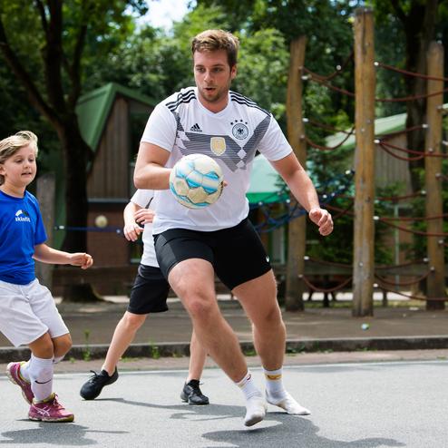 Ein Junge in blauem Shirt und ein Mann in wei&szlig;em Shirt spielen Fu&szlig;ball auf einem Platz mit B&auml;umen im Hintergrund.