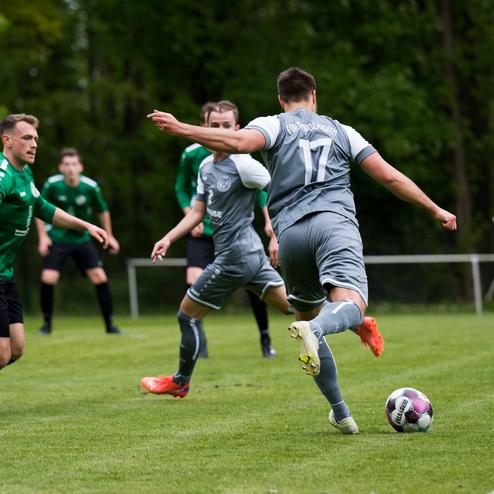 Ein Fu&szlig;ballspieler in grauem Trikot schie&szlig;t den Ball, w&auml;hrend andere Spieler in gr&uuml;nen Trikots reagieren.