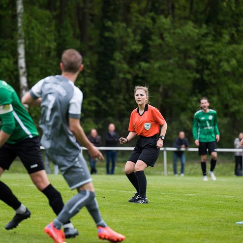 Zwei Fu&szlig;ballspieler in grauen Trikots und ein Schiedsrichter in Orange beobachten einen Ball auf dem Spielfeld.