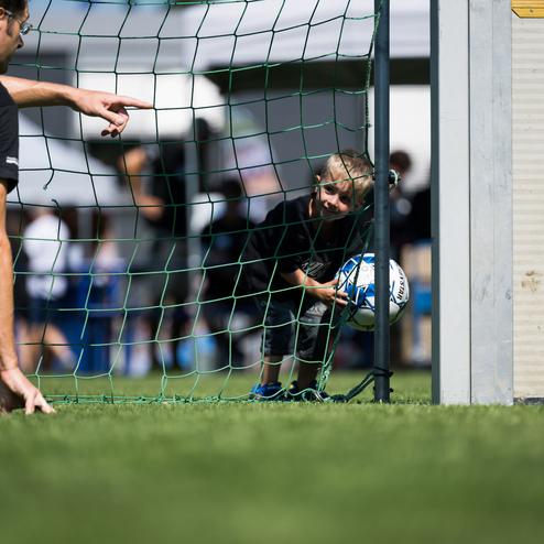 Ein Junge versteckt sich hinter einem Fu&szlig;balltor und h&auml;lt einen Fu&szlig;ball, w&auml;hrend ein Erwachsener auf ihn zeigt.