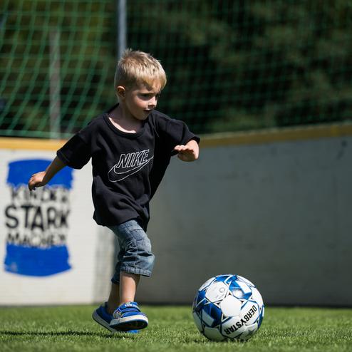 Ein Junge in einem schwarzen T-Shirt kickt einen Fu&szlig;ball auf einem gr&uuml;nen Spielfeld mit einem Tor im Hintergrund.
