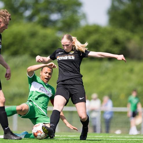 Zwei Fu&szlig;ballspieler im Zweikampf auf dem Platz, eine Spielerin springt &uuml;ber einen liegenden Spieler in gr&uuml;ner Ausr&uuml;stung.