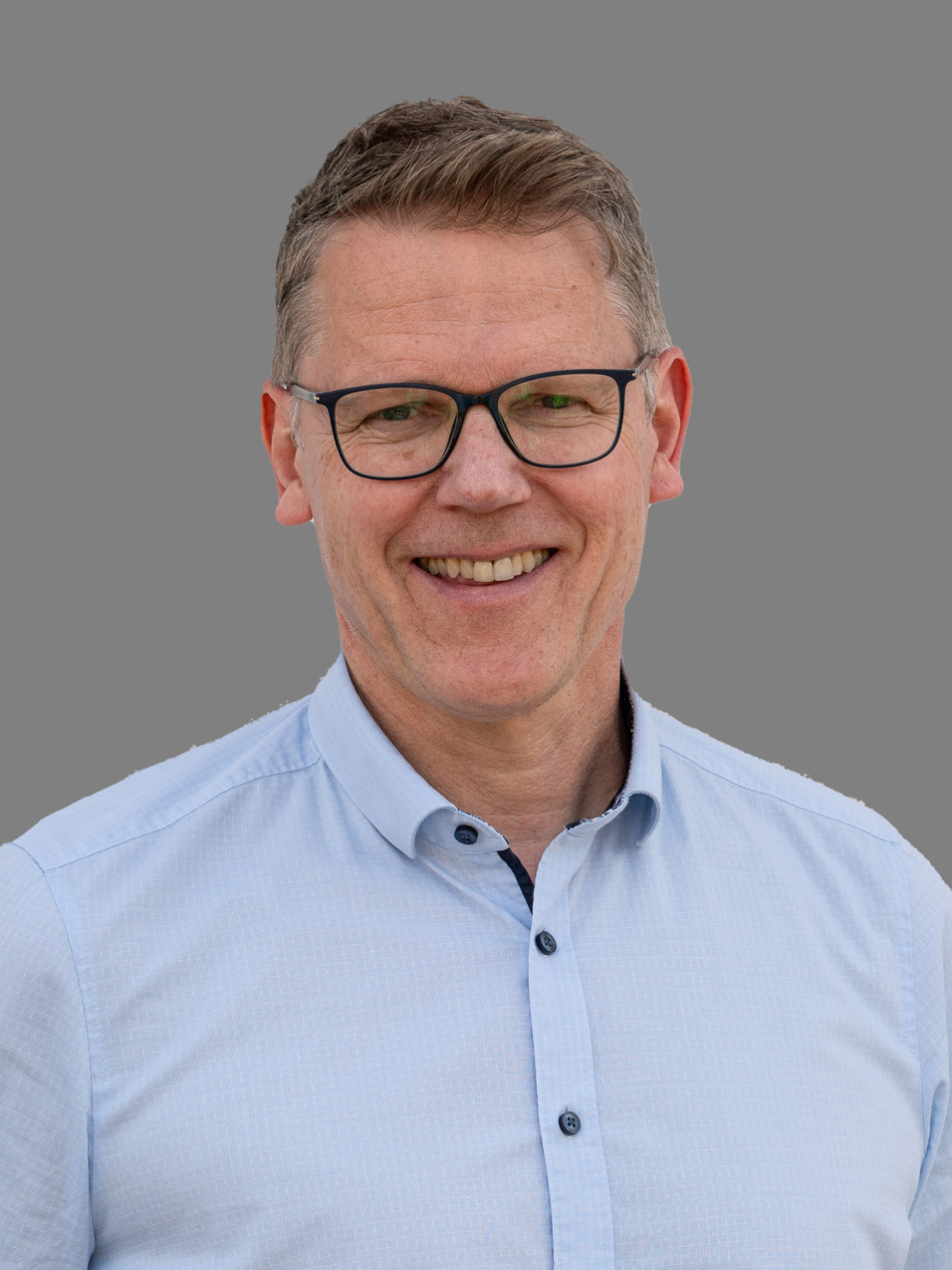 Ein l&auml;chelnder Mann mit Brille tr&auml;gt ein hellblaues Hemd und steht vor einem grauen Hintergrund.