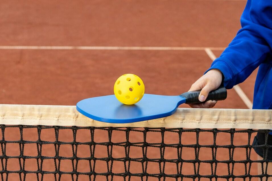 Ein blauer Schl&auml;ger mit einer gelben Pickleball-Kugel liegt auf einem Netz einer Tennisplatz-Bahnen mit rotem Belag.
