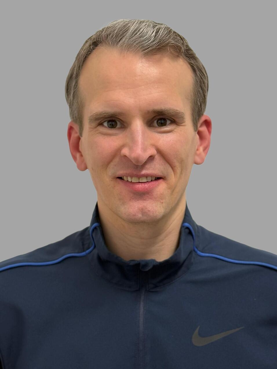 Portr&auml;t eines l&auml;chelnden Mannes in dunkler Sportjacke mit blauem Akzent vor einfarbig grauem Hintergrund.