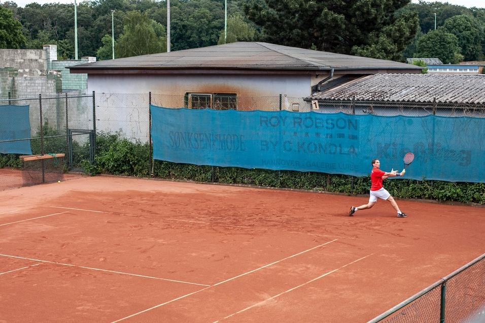 Tennisplatz mit einem Spieler in rotem Shirt, der einen Ball mit einem Schl&auml;ger auf rotem Sandcourt anschl&auml;gt.