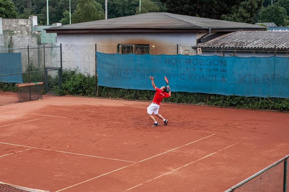 Ein Tennisspieler in roten Shirt beim Aufschlag auf einem roten Sandplatz, im Hintergrund eine blaue Werbebande.