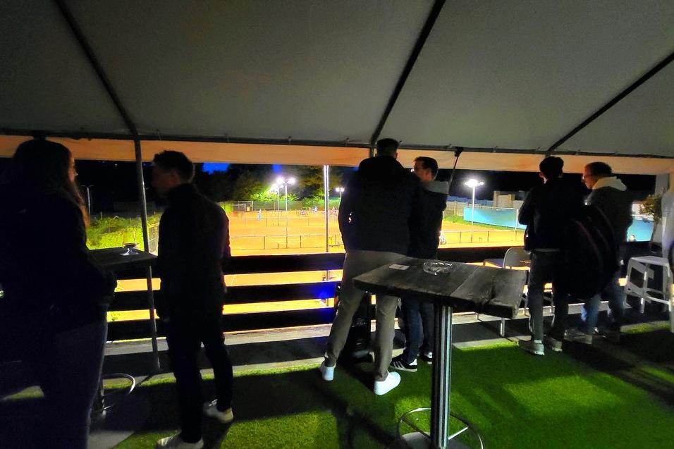 Gruppe von Menschen steht unter einem Zelt und beobachtet ein beleuchtetes Tennisfeld bei Nacht.