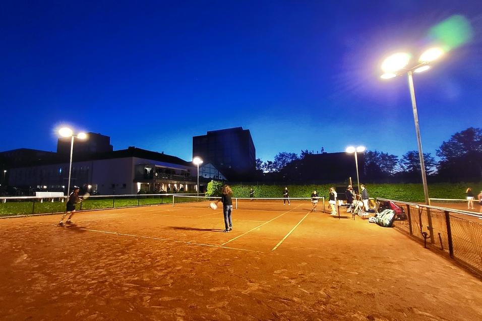 Tennisspieler auf einem beleuchteten Sandplatz bei D&auml;mmerung, umgeben von Geb&auml;uden und Gr&uuml;nfl&auml;chen.
