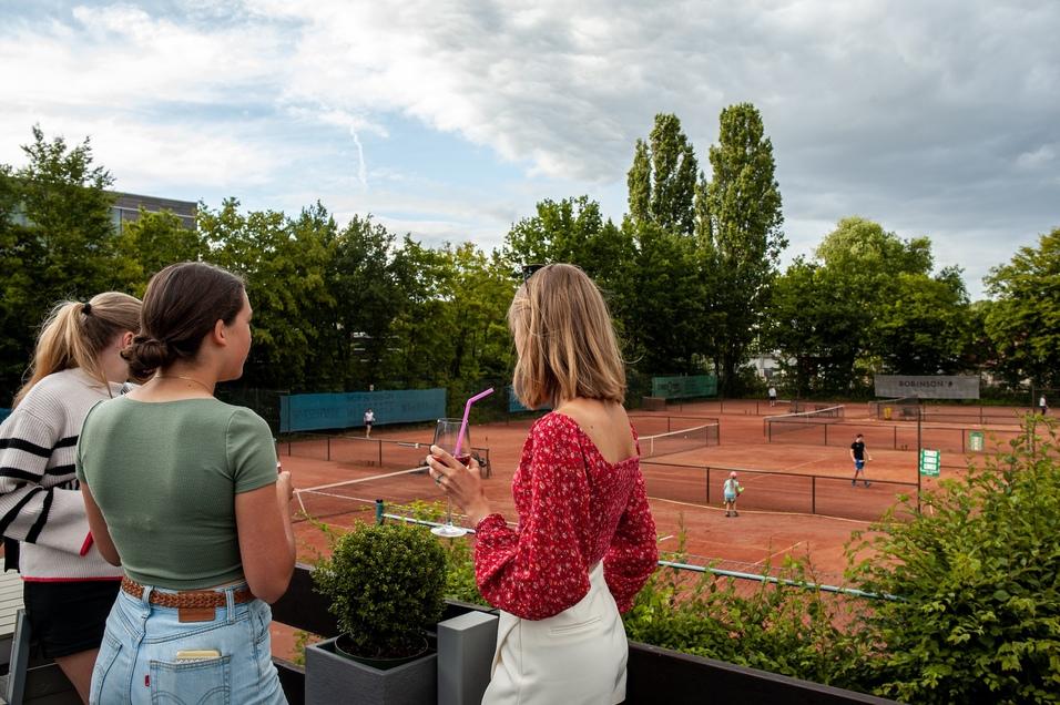 Drei Frauen beobachten ein Tennisspiel auf roten Sandpl&auml;tzen, umgeben von B&auml;umen und einem bew&ouml;lkten Himmel.