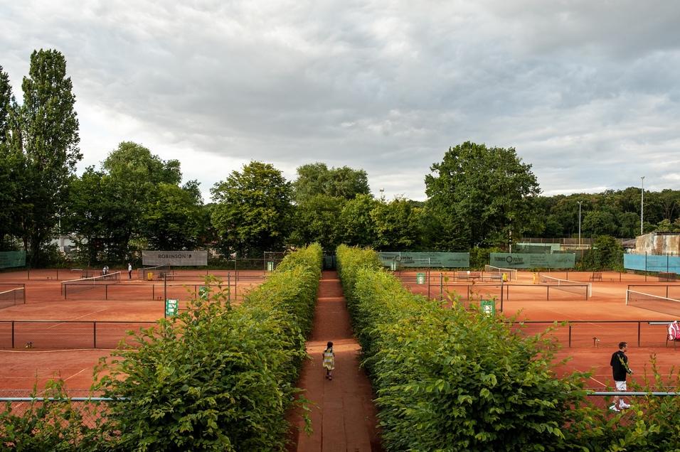Blick auf einen Tennisplatz mit mehreren Spielfeldern, umgeben von gr&uuml;nen Hecken und B&auml;umen, zwei Personen sind sichtbar.