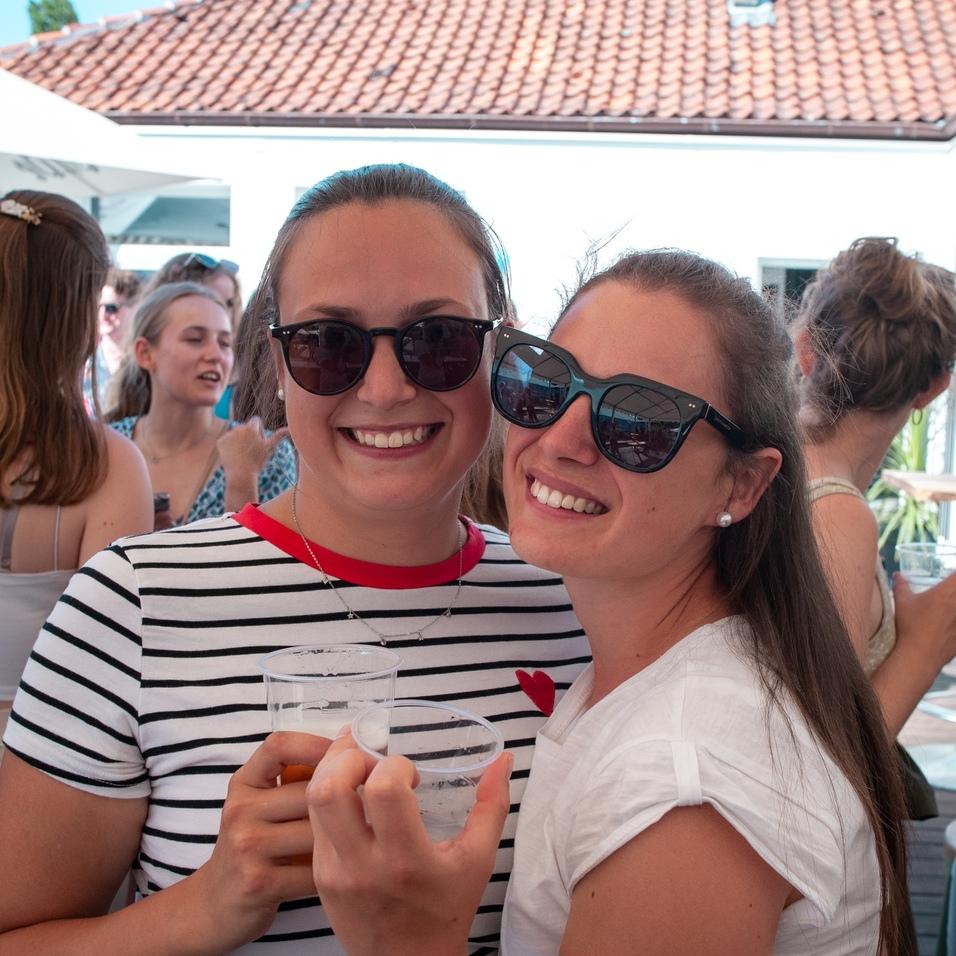 Zwei Frauen lächeln, halten Plastikbecher mit Getränken und tragen Sonnenbrillen, umgeben von Feiernden im Hintergrund.
