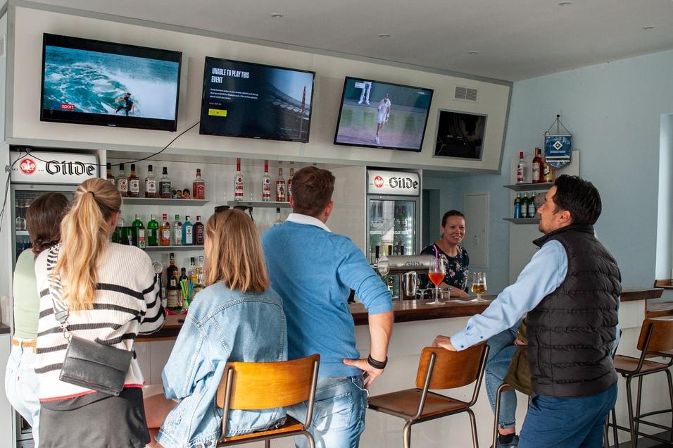 Gruppe von G&auml;sten an einer Bar, mit Getr&auml;nken, w&auml;hrend sie Sport&uuml;bertragungen auf Bildschirmen verfolgen.