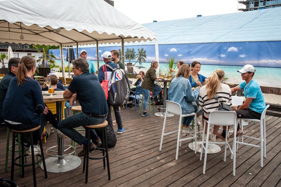 Menschen sitzen in einem Strandcaf&eacute; und genie&szlig;en Getr&auml;nke sowie Snacks unter einem Sonnenzelt mit Palmenkuppel im Hintergrund.