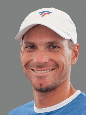 L&auml;chelnder Mann mit kurz rasierter Frisur und wei&szlig;er Kappe vor grauem Hintergrund, tr&auml;gt ein blaues Shirt.