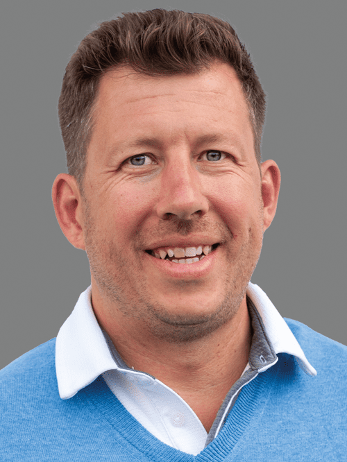 Portr&auml;t eines Mannes mit kurzen Haaren, blauem Pullover und wei&szlig;em Hemdkragen, vor grauem Hintergrund.