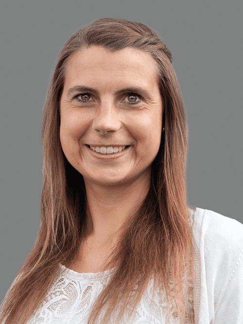 Portr&auml;t einer l&auml;chelnden Frau mit langen, braunen Haaren und einem wei&szlig;en Oberteil vor grauem Hintergrund.