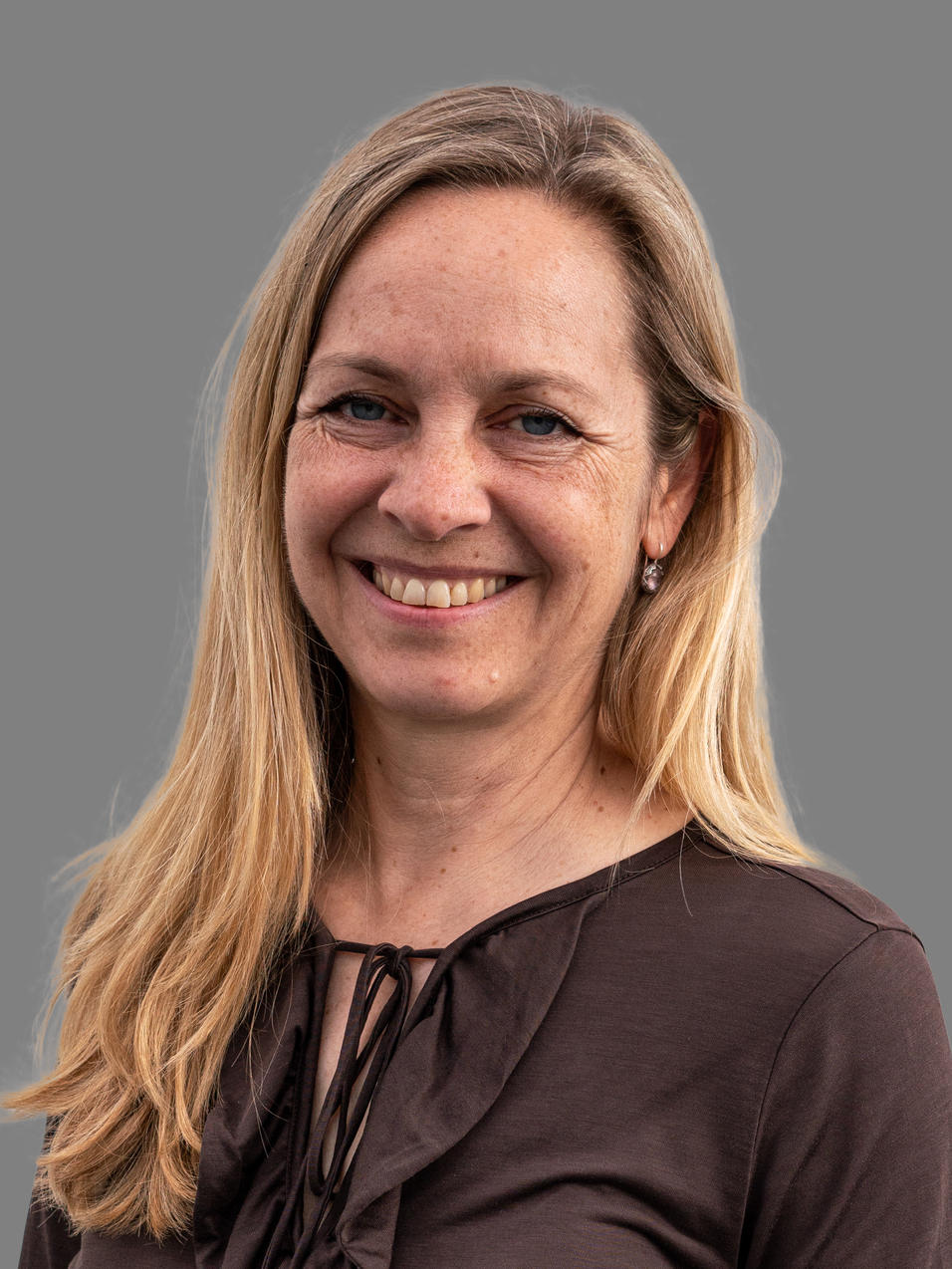 L&auml;chelnde Frau mit langen, blonden Haaren, die ein dunkelbraunes Oberteil tr&auml;gt, vor grauem Hintergrund.