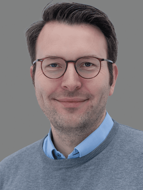 Portr&auml;t eines Mannes mit Brille, l&auml;chelnd, in einem grauen Pullover und blauem Hemd, vor grauem Hintergrund.