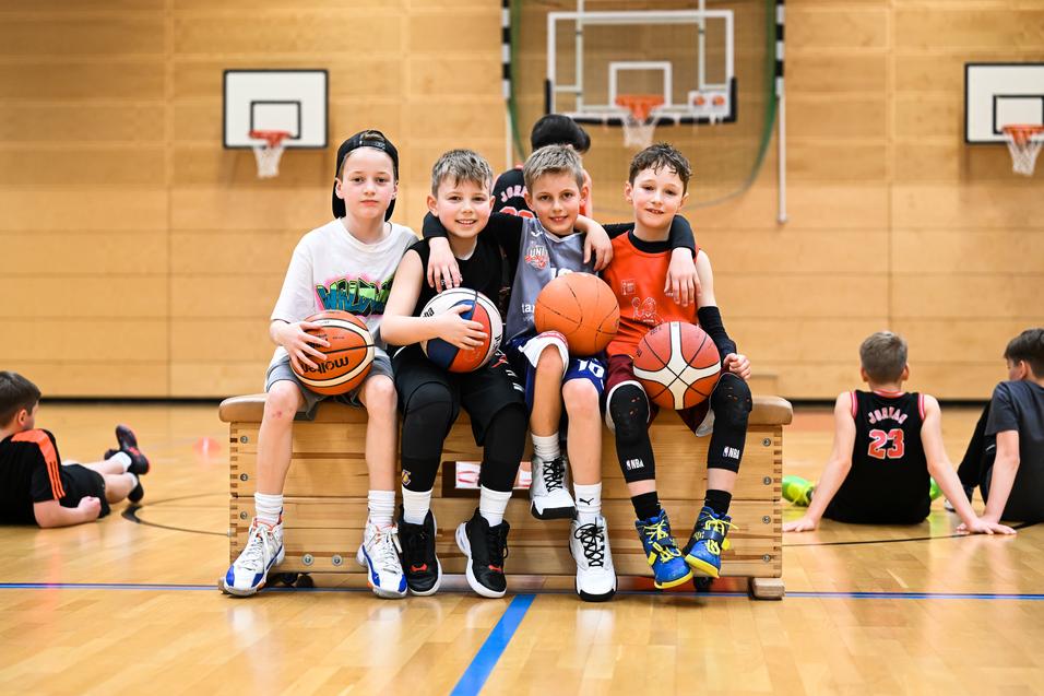 Vier Kinder sitzen auf einer Bank in der Turnhalle, halten Basketballs und lachen. Im Hintergrund sind weitere Kinder aktiv.