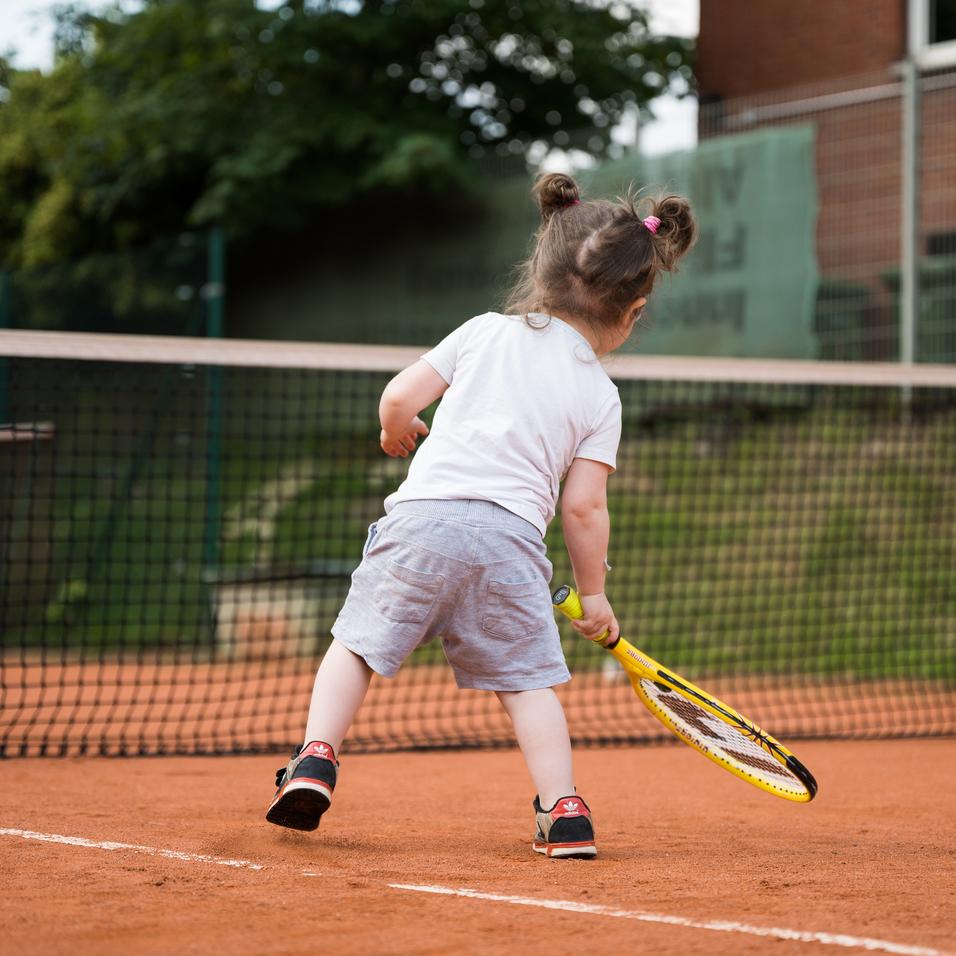 Kind mit Zöpfen schlägt Tennisball auf einem Sandplatz, während es sich auf einen Schlag vorbereitet.