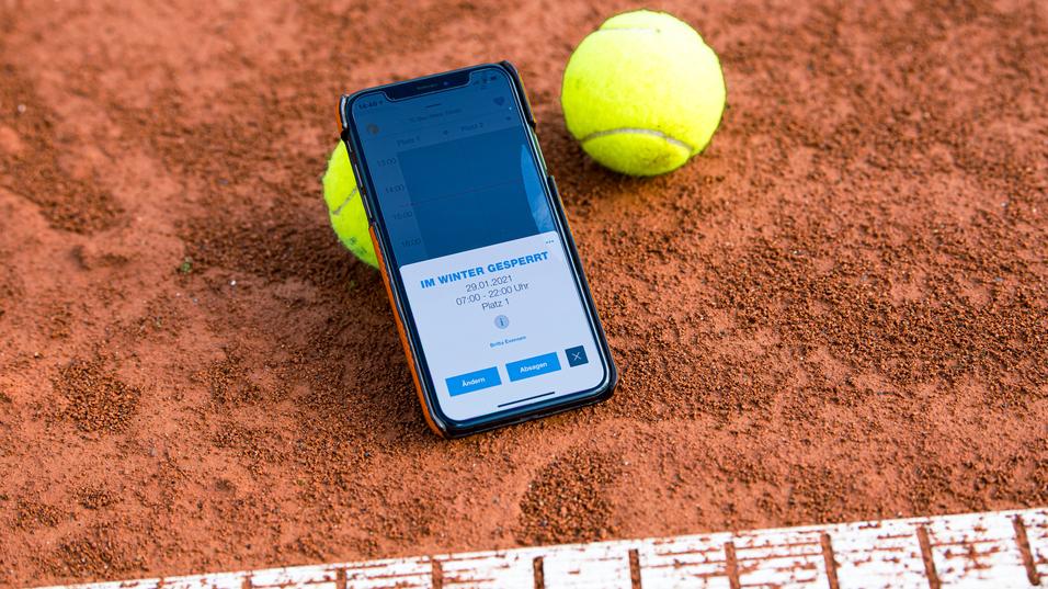 Smartphone mit Buchungsinfo liegt auf einem Tennisplatz, neben zwei gelben Tennisbällen auf rotem Sand.