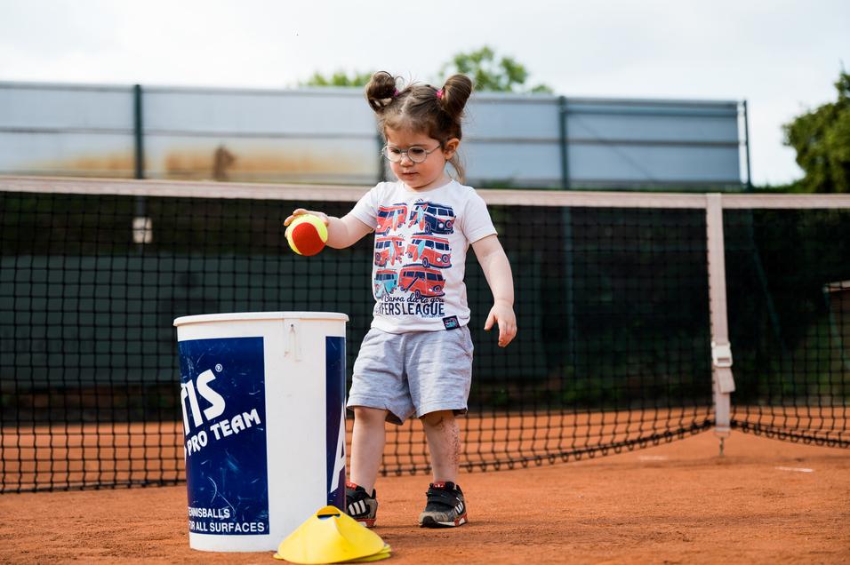 Kleines Kind mit Brille steht neben einem Eimer und hält einen Tennisball auf einem Tennisplatz.