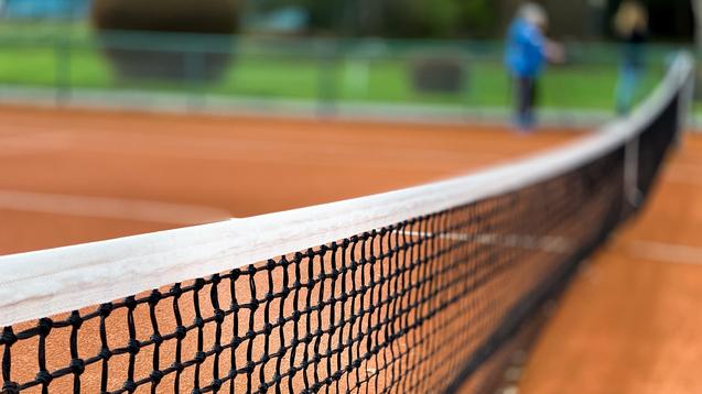 Nahaufnahme eines Tennisnetzes auf einem roten Sandplatz, Hintergrund unscharf mit grünen Bäumen.